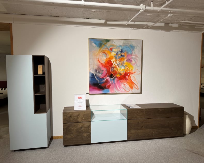 Die MAB Wohnkombination Sona mit Regal und Glasvitrine wird in einem Ausstellungsraum mit einem abstrakten, farbenfrohen Kunstwerk darüber präsentiert.