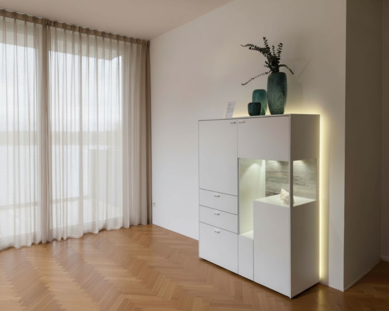 Minimalistisches Wohnzimmer mit transparenten Vorhängen, Holzfußboden und dem modernen Venjakob Highboard Sentino 3000 in Weiß, dekoriert mit stilvollen Vasen darauf.