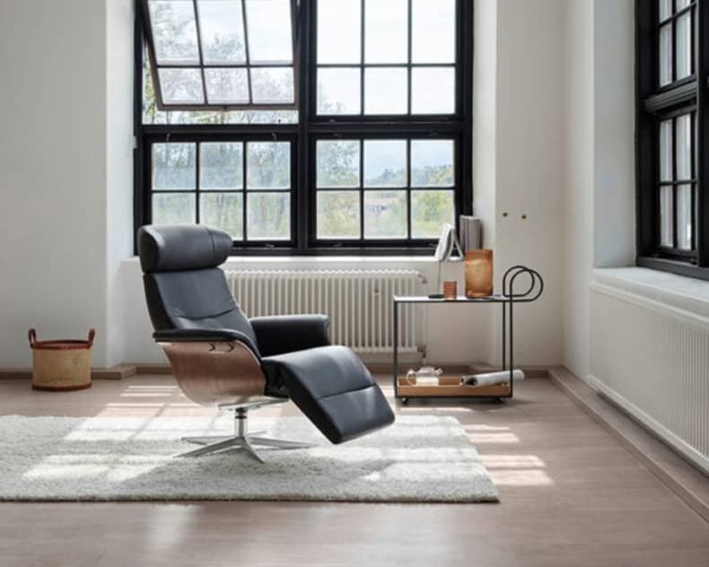 Ein minimalistischer Raum ist mit dem schwarzen Loungesessel SALE 25 Sessel Time Out, einem Beistelltisch und großen Gitterfenstern ausgestattet. Licht strömt herein und erhellt den Raum.