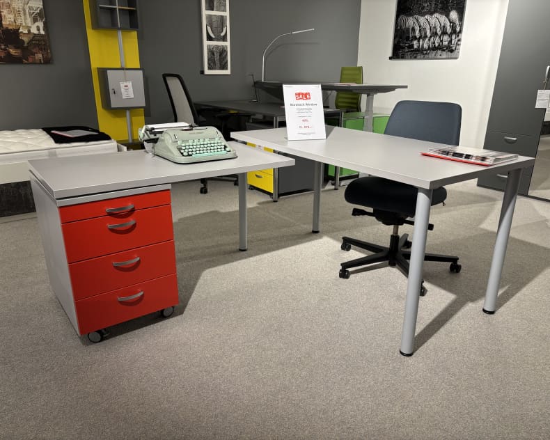 Büroeinrichtung bestehend aus Büromöbeln mit 3 Boxen Window, einem grauen Schreibtisch, einem roten Schubladenelement, Schreibmaschine, Bürostuhl und Dekor im Hintergrund.