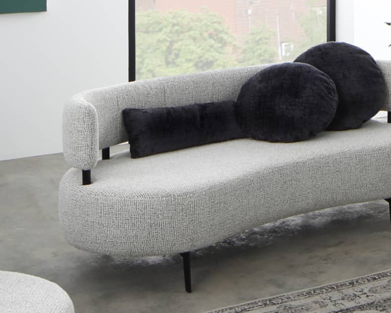 In einem hellen Raum mit großen Fenstern verfügt das Ponsel Sofa Organic in modernem Grau über schwarze runde und zylindrische Kissen, ergänzt durch einen Pflanzenkorb.