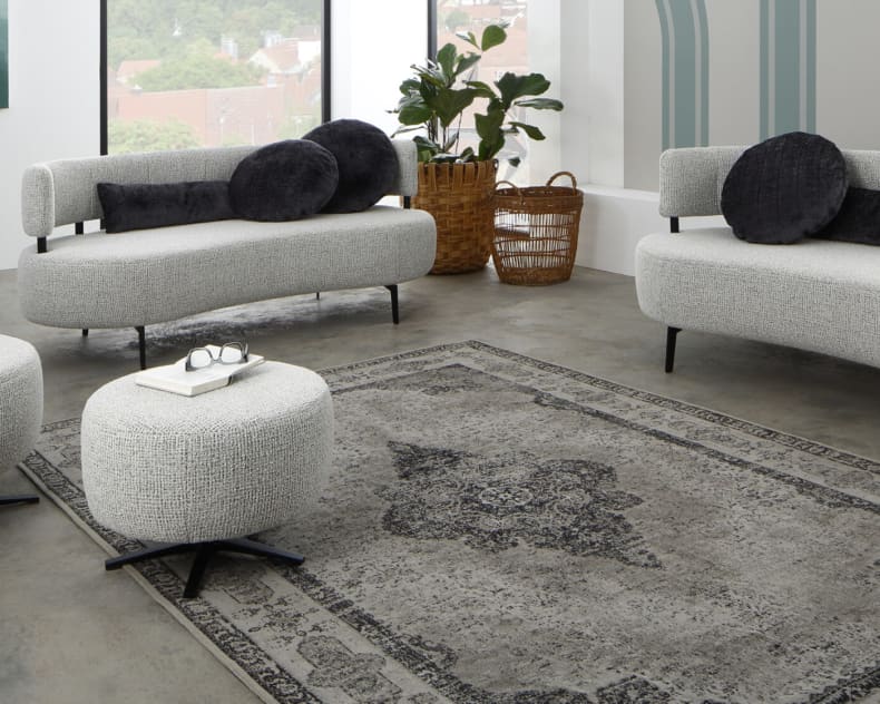Modernes Wohnzimmer mit Ponsel Sofa Organic, rundem Ottoman und dekorativem Teppich. Große Fenster und Zimmerpflanzen sorgen für Helligkeit.