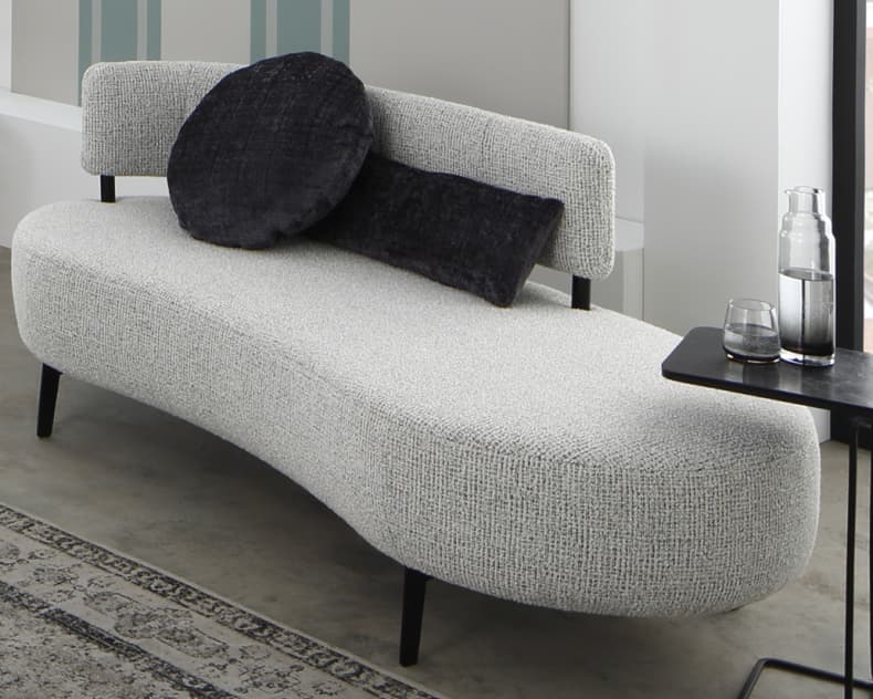 Ein geschwungenes graues Sofa von Ponsel Recamiere Organic mit zwei dunklen, runden Kissen steht neben einem schwarzen Beistelltisch, auf dem eine Glaskanne und ein Glas stehen.