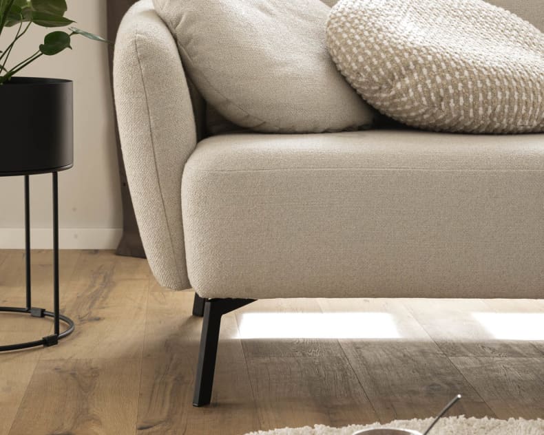 Das Candy Sofa Nimba in Beige ist mit Kissen kombiniert und steht neben einer Topfpflanze auf einem Holzboden. Daneben steht ein runder Tisch mit einer Tasse, alles beleuchtet vom natürlichen Licht des Fensters.