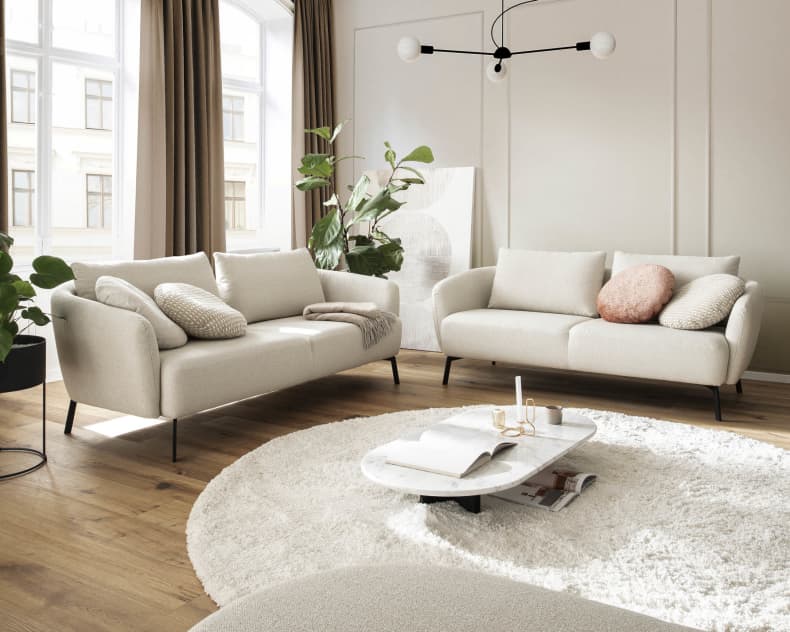 Ein gemütliches Wohnzimmer mit Candy Sofa Nimba, einem hellen Sofa, einem runden Teppich, einem Marmor-Couchtisch und Pflanzen an großen Fenstern.