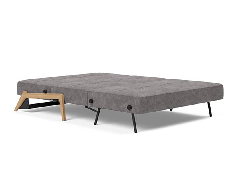 Das Innovation Schlafsofa Cubed 140 ist ein modernes graues Futonsofa mit schwarzen Metallbeinen und Armlehnen aus Holz, hier in der flachen Position gezeigt.