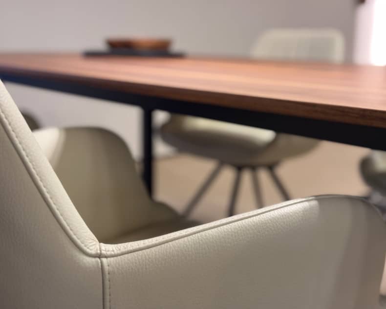 Nahaufnahme eines Willisau Stuhl Loreo in Beige neben einem Holztisch, mit unscharfen Stühlen und einer Schüssel im Hintergrund.