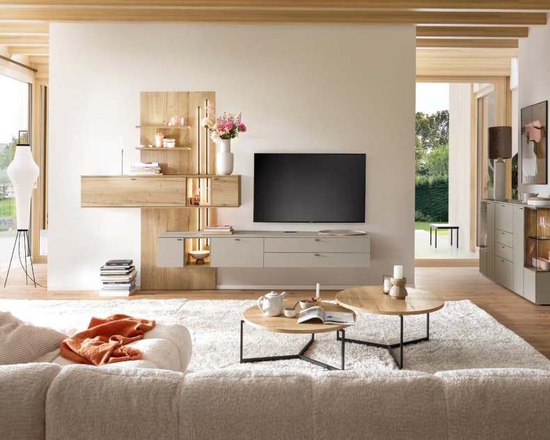 Das Venjakob Wohnprogramm Vanya bietet eine moderne Wohnzimmereinrichtung mit neutralen Tönen, einem großen Fernseher, Holzregalen und einem gemütlichen beigen Sofa, das durch einen stilvollen Teppich ergänzt wird.