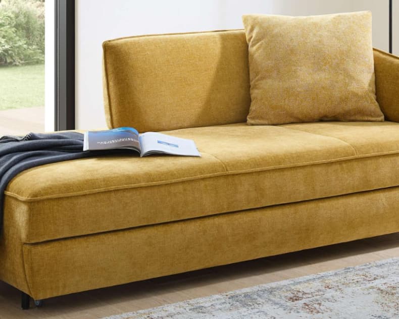 Das Bali Bettsofa Ilka in Gelb steht zusammen mit einem passenden Kissen, einer dunklen Decke und einem offenen Buch in einem hellen Wohnzimmer.