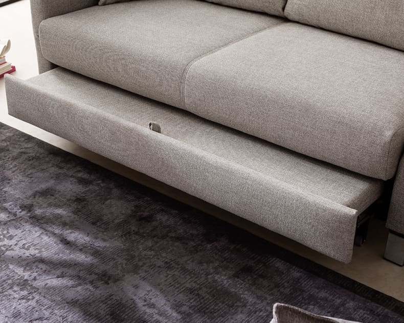 Das Bali Bettsofa Tilda, ein graues Sofa mit ausziehbarem Bettgestell, steht neben einem dunklen Strukturteppich und gestapelten Büchern.
