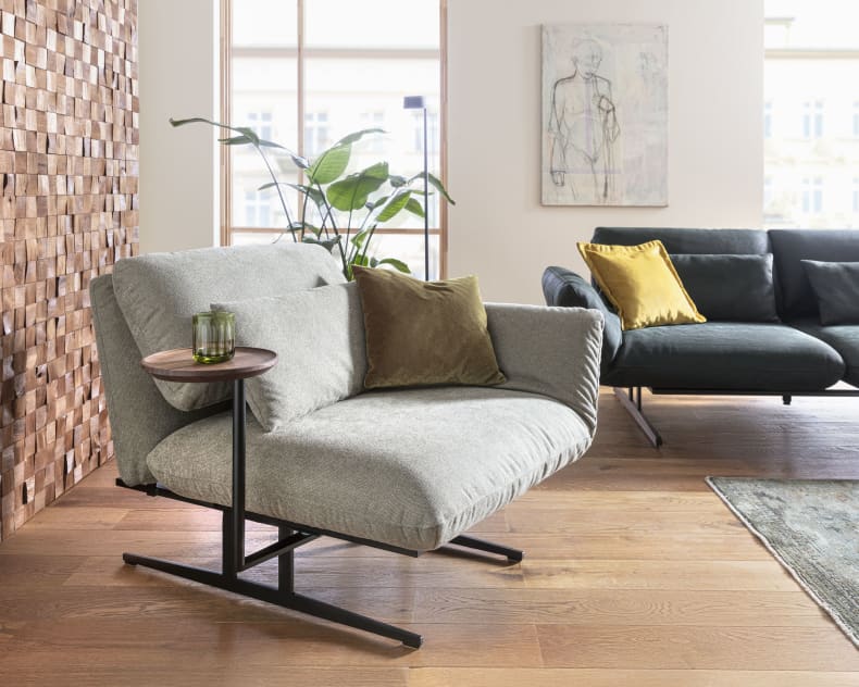 Ein modernes Wohnzimmer mit dem grauen Sessel Signet Sessel Messina, Beistelltisch, Kissen, Pflanze und Sofa auf einem Holzboden.