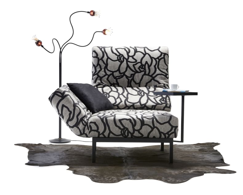 Das Signet Sofa Chimba zeichnet sich durch ein modernes Design mit einem auffälligen Blumenmuster aus, gepaart mit einem schwarzen Kissen, einem Beistelltisch, einer Stehlampe und einem Kuhfellteppich auf weißem Hintergrund.