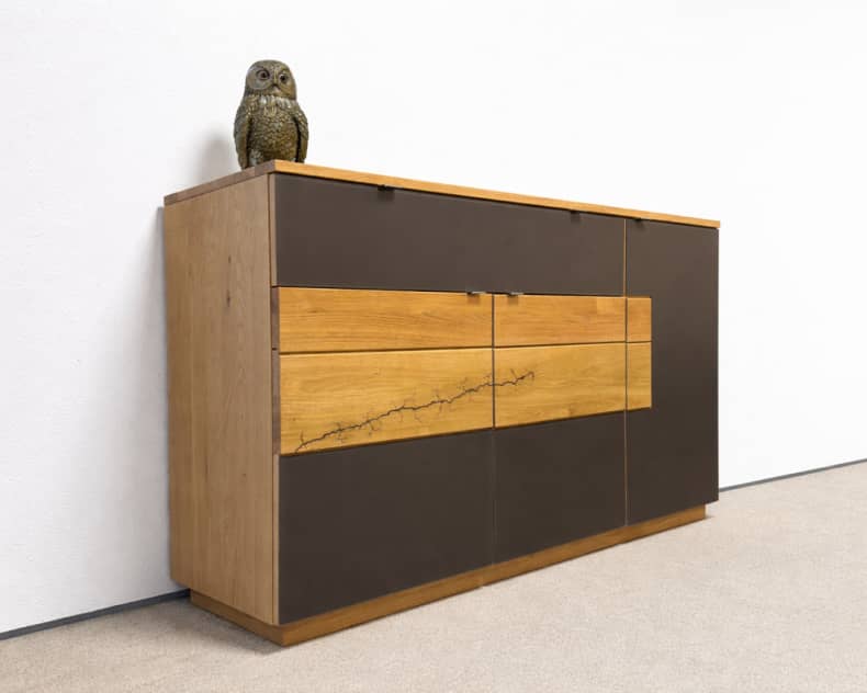 Das Wimmer Sideboard Signatura zeichnet sich durch sein modernes Holzdesign mit dunklen Paneelen aus und wird von einer Eulenskulptur gekrönt, die vor einer weißen Wand steht.