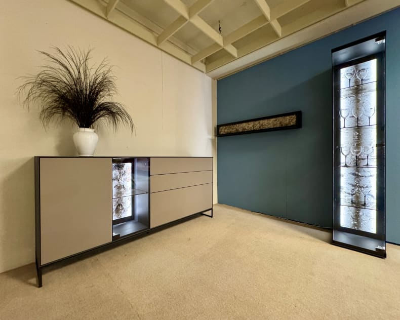 Modernes, minimalistisches Zimmer mit dem beigen Hartmann Wohnprogramm Alva Sideboard, einer hohen Glasvitrine und einer Vase mit hohem schwarzem Gras.