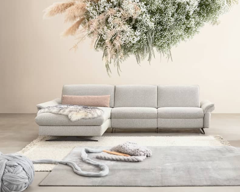 Modernes Wohnzimmer mit dem grauen Sofa Himolla Polstergruppe Spektra 1178, einem weichen Teppich, Akzenten aus Grobstrick und einem hängenden Blumenarrangement darüber.