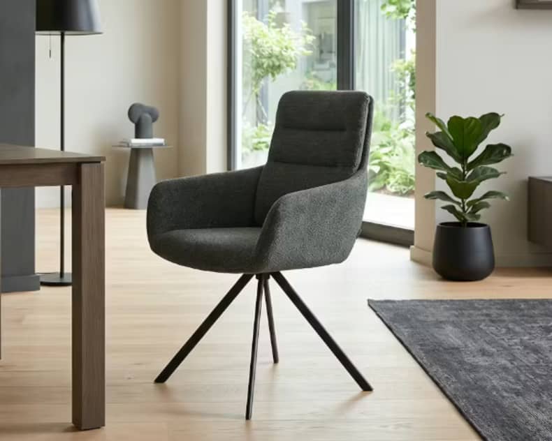 Der Venjakob Stuhl Felia ist ein moderner grauer Polsterstuhl mit angewinkelten Beinen, der in einem hellen, minimalistischen Wohnbereich mit Pflanzen und großen Fenstern gezeigt wird.