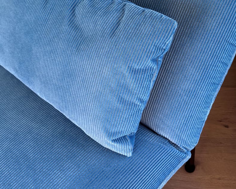 Das blaue Kordkissen Liege Roro 98 liegt auf einem passenden Sofa, im Hintergrund ist ein Holzboden zu sehen.