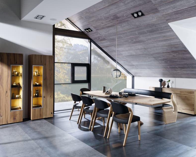 Modernes Esszimmer mit Holzmöbeln, Voglauer Stuhl 35 in Schwarz und großen Fenstern unter einer schrägen Decke.