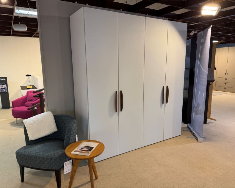 Der Jutzler Panorama-Falttürenschrank mit braunen Griffen steht neben einem grauen Stuhl und einem kleinen runden Holztisch im Ausstellungsraum.