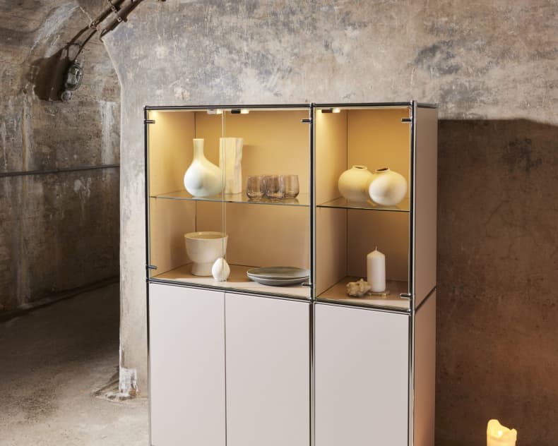 Die moderne Vitrine „MAB“ mit Keramikobjekten steht vor einer strukturierten Wand, flankiert von brennenden Kerzen auf dem Boden.