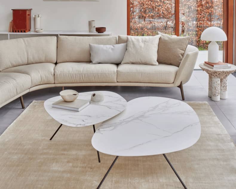 Ein modernes Wohnzimmer mit einem beigen, geschwungenen Sofa, Leolux-Beistelltischen „Liliom“ aus Marmor, einem hellen Teppich und großen Fenstern, durch die man die herbstlichen Bäume draußen sehen kann.