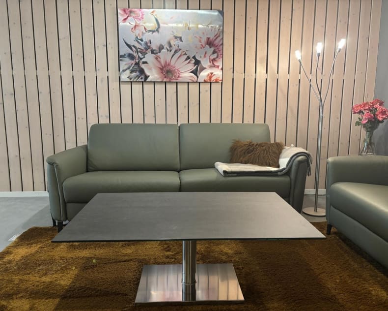 Ein modernes Wohnzimmer mit dem grünen Sofa „Ravenna“ aus der PM Oelsa Polstergruppe, einem schwarzen Tisch, einem Kunstwerk mit Blumenmotiv, einer Stehlampe und einem braunen Teppich.