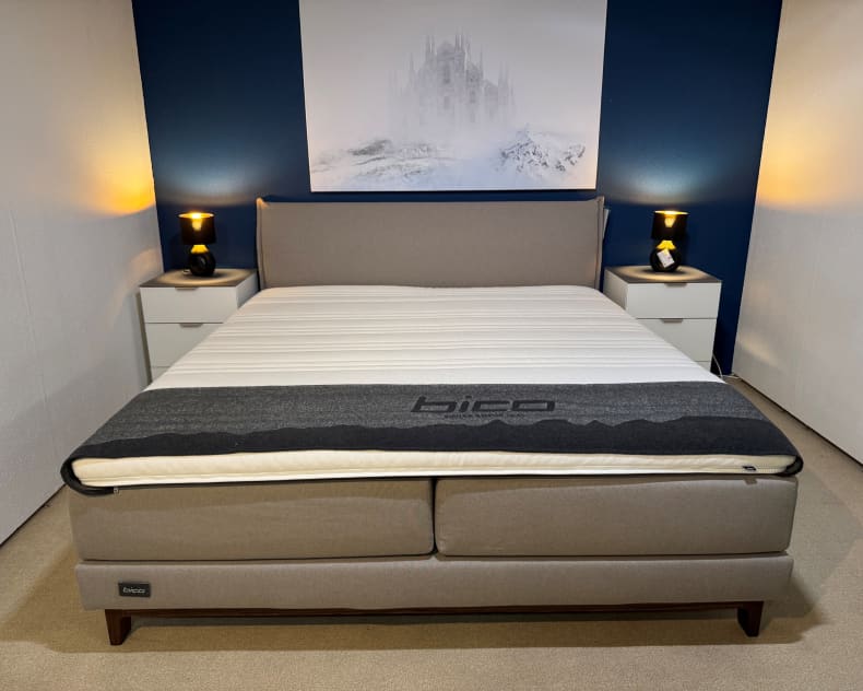 Vor einer blauen Akzentwand steht ein gepflegtes Bico-Boxspringbett mit grauem Kopfteil, flankiert von zwei Nachttischen und Lampen.