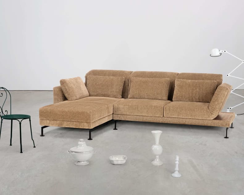 Brühl-Sofa „Moule Small“ in Hellbraun, schwarzer Metallstuhl, weiße Lampe und verschiedene weiße Dekoartikel auf einem grauen Boden.