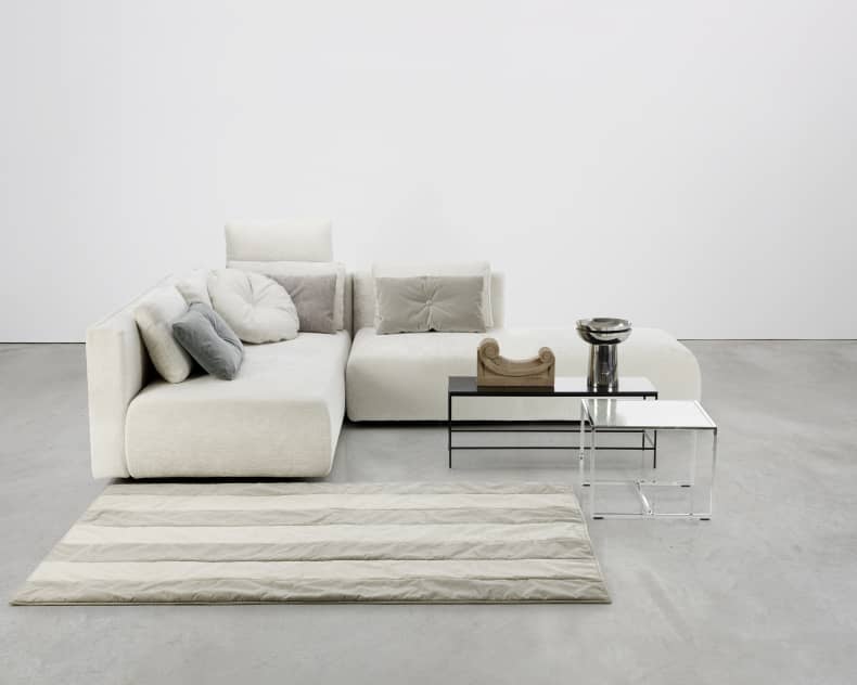 Le canapé fonction Brühl „Piano Small“ en version moderne couleur crème, équipé de coussins neutres, combiné avec deux tables d'appoint et placé sur un tapis beige à rayures sur un sol gris.