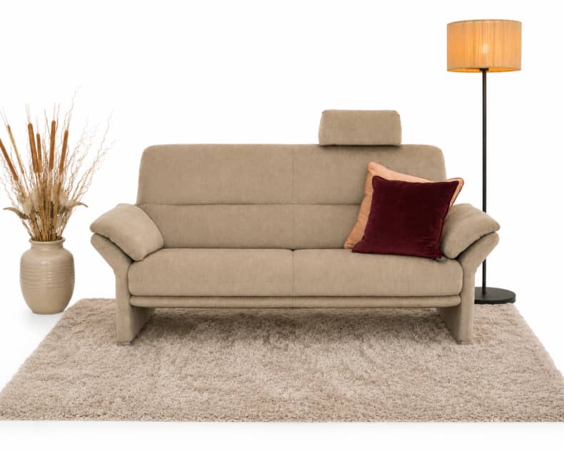 Horst-Sofa „Grimsel“ (2,5-Sitzer) mit einem roten Kissen, einer Lampe und einer Vase mit getrocknetem Gras auf einem beigen Teppich vor weißem Hintergrund.