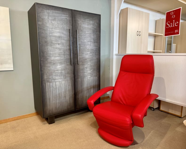 Neben dem „Sprenger Schrank 2-türig“, einem hohen, dunklen Schrank, steht ein roter Ledersessel in einem Ausstellungsraum, im Hintergrund ist ein rotes Verkaufsschild zu sehen.