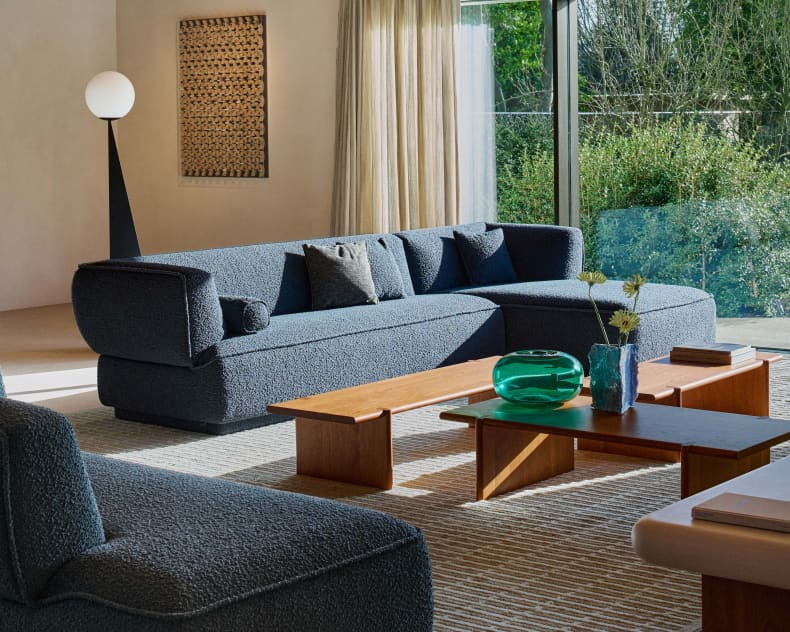 Un salon moderne avec le canapé bleu Leolux « Loya », complété par des tables en bois, une lampe sur pied, de grandes fenêtres et une vue sur la verdure.