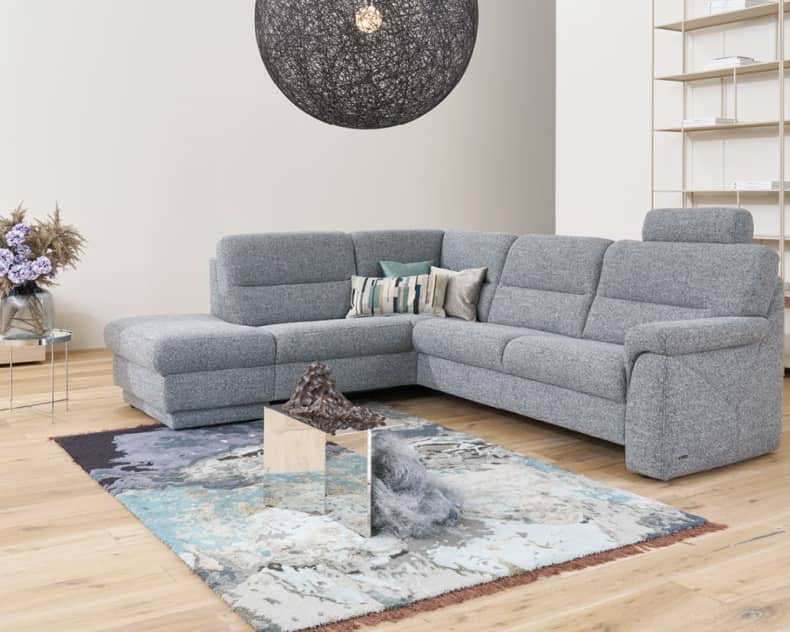 Das Himolla-Sofa 1101 in einem hellen Wohnzimmer mit einem Teppich, einer modernen Lampe und Regalen an der Wand.