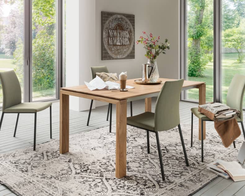 Modernes Esszimmer mit dem Holztisch Sit Mobilia Tisch Merkur Allegro, vier grünen Stühlen und großen Fenstern mit Blick auf einen üppigen Garten.