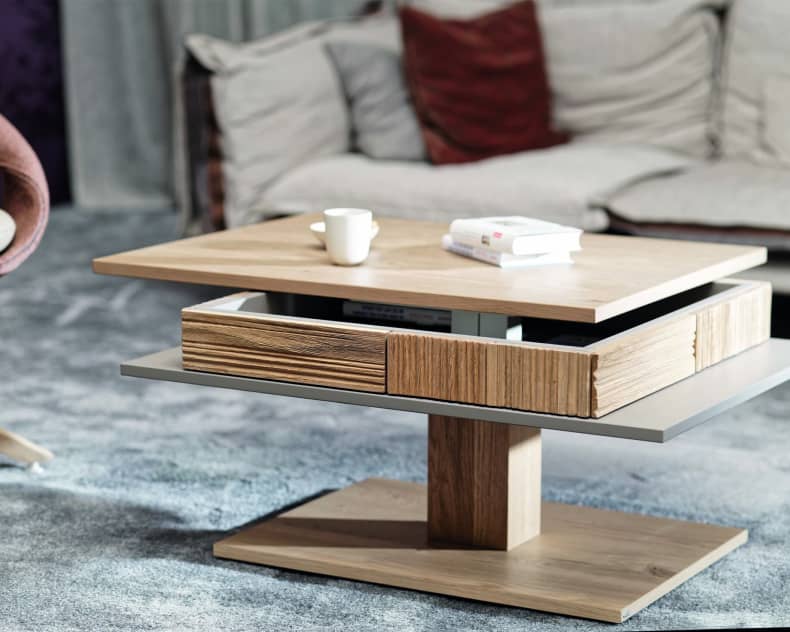 Der Voglauer Salontisch LAC77 ist ein moderner Couchtisch aus Holz mit drehbaren Etagen, auf dem Bücher und eine Tasse in einem gemütlichen Wohnzimmer stehen.