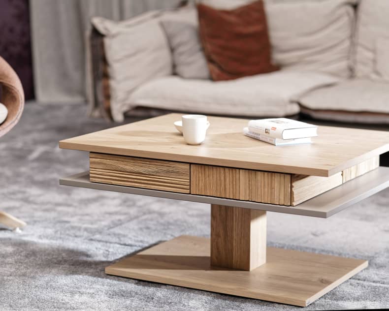 Der Voglauer Salontisch LAC77, ein moderner Couchtisch aus Holz mit Büchern und einer Tasse, steht in einer gemütlichen Wohnlandschaft mit einem Sofa im Hintergrund.