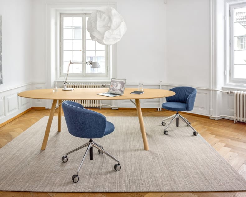 Ein minimalistisches Büro mit dem Holztisch „Esstisch Akio Oval“ von Girsberger, zwei blauen Stühlen, einem Laptop, einer Lampe und einem großen Fenster, durch das Tageslicht hereinströmt.