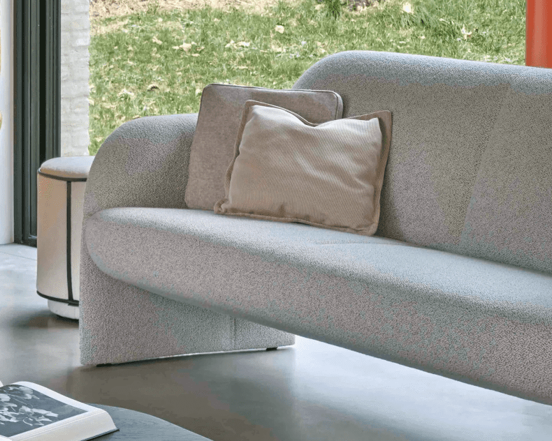 Leolux Sofa Modius in Hellgrau mit zwei beigen Kissen, positioniert an einem Fenster mit Blick auf eine grüne Wiese.