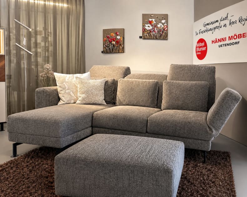 Ein modernes „Brühl Sofa Moule Small“ in Grau mit Kissen steht auf einem braunen Teppich, ergänzt durch Wandbilder und ein dekoratives Schild im Hintergrund.