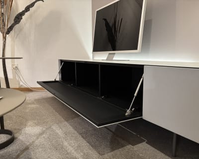 Modernes Wohnzimmer mit einem Flachbildfernseher auf einem Büromöbel mit 3 Boxenfenstern, einem eleganten weißen Schrank mit offenem Stauraum, alles auf einem grauen Teppich.