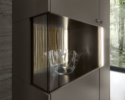 Die SALE 25 Wohnkombination Linee, eine Glasschale, präsentiert in einem modernen Schrank mit Glastüren und beleuchtetem Innenraum, vor dem Hintergrund eines Waldblicks.