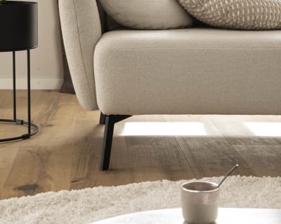 Das Candy Sofa Nimba in Beige ist mit Kissen kombiniert und steht neben einer Topfpflanze auf einem Holzboden. Daneben steht ein runder Tisch mit einer Tasse, alles beleuchtet vom natürlichen Licht des Fensters.