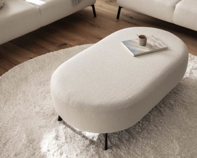 Das Candy Sofa Nimba dient als perfekte Kulisse für einen ovalen beigen Ottomanen, auf dem ein Buch und eine Tasse liegen, die alle auf einem runden beigen Teppich im Wohnzimmer ruhen.