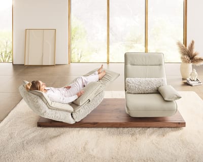Une personne se détend sur les fonctions de Koinor de l&#39;Evino, une chaise de repos réglable et douce, dans une pièce lumineuse avec de grandes fenêtres et un décor minimaliste.