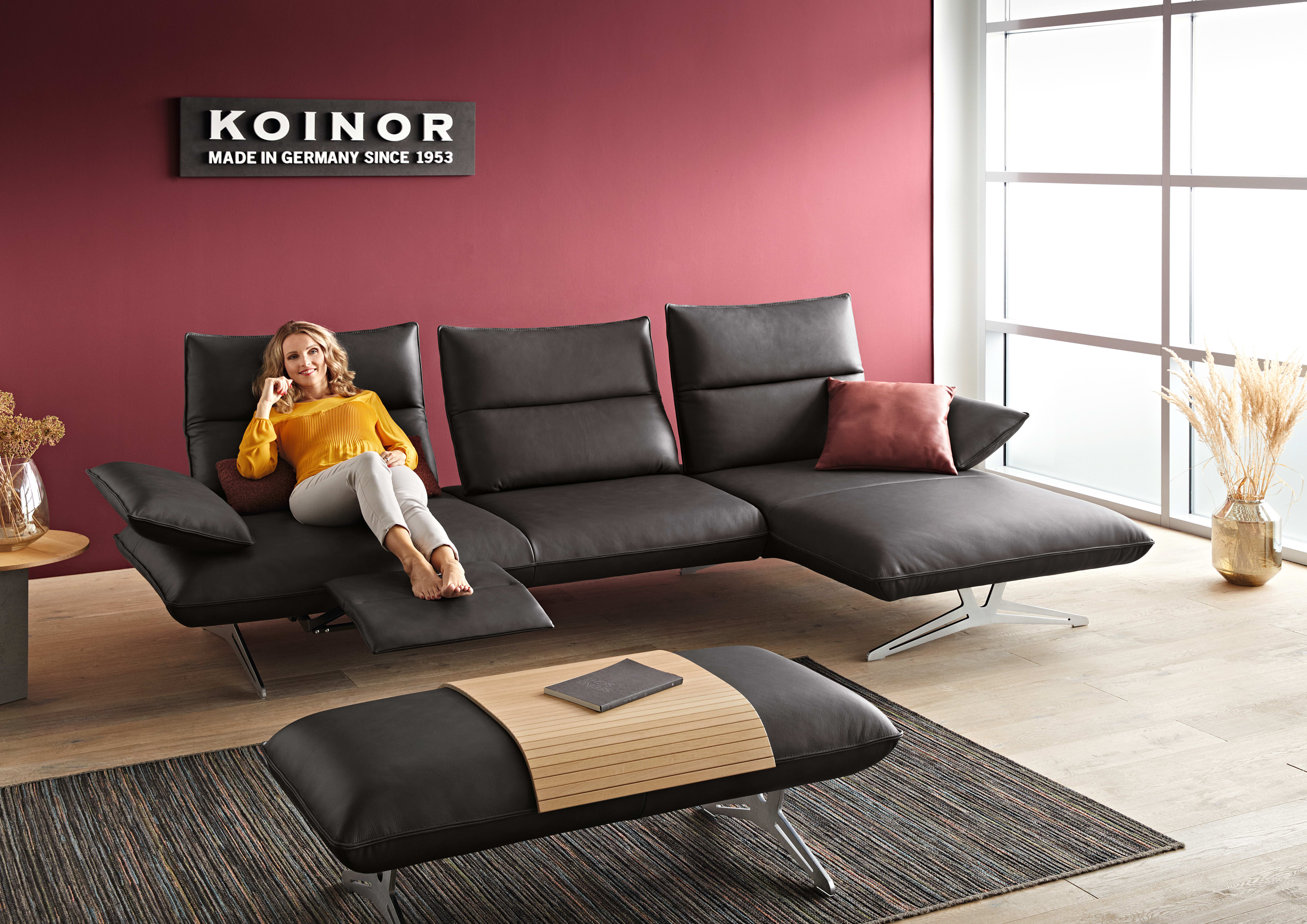 Une femme se détend sur le canapé d&#39;angle noir Caio de Koinor Polstergruppe dans un salon élégant agrémenté d&#39;un mur d&#39;accent violet.