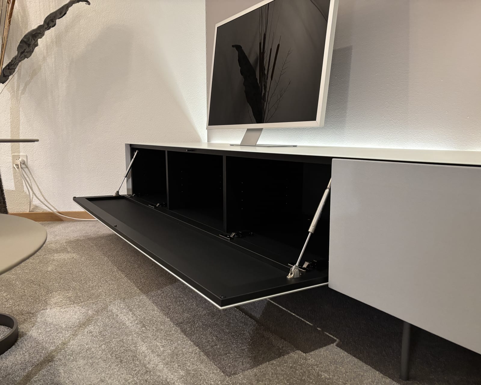 Modernes Wohnzimmer mit einem Flachbildfernseher auf einem Büromöbel mit 3 Boxenfenstern, einem eleganten weißen Schrank mit offenem Stauraum, alles auf einem grauen Teppich.