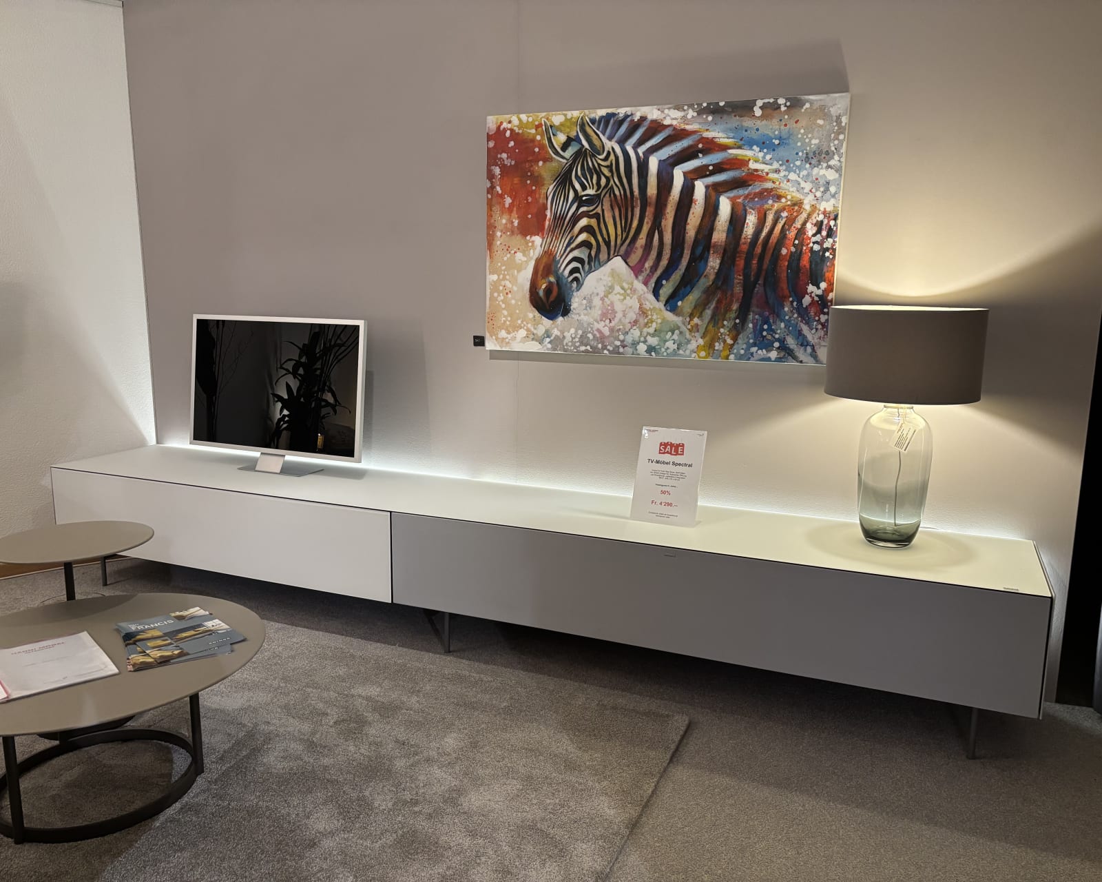 Moderne Wohnzimmereinrichtung mit Fernseher, Zebragemälde, Lampe und runden Couchtischen auf einem grauen Teppich mit Büromöbeln mit 3 Boxenfenstern.