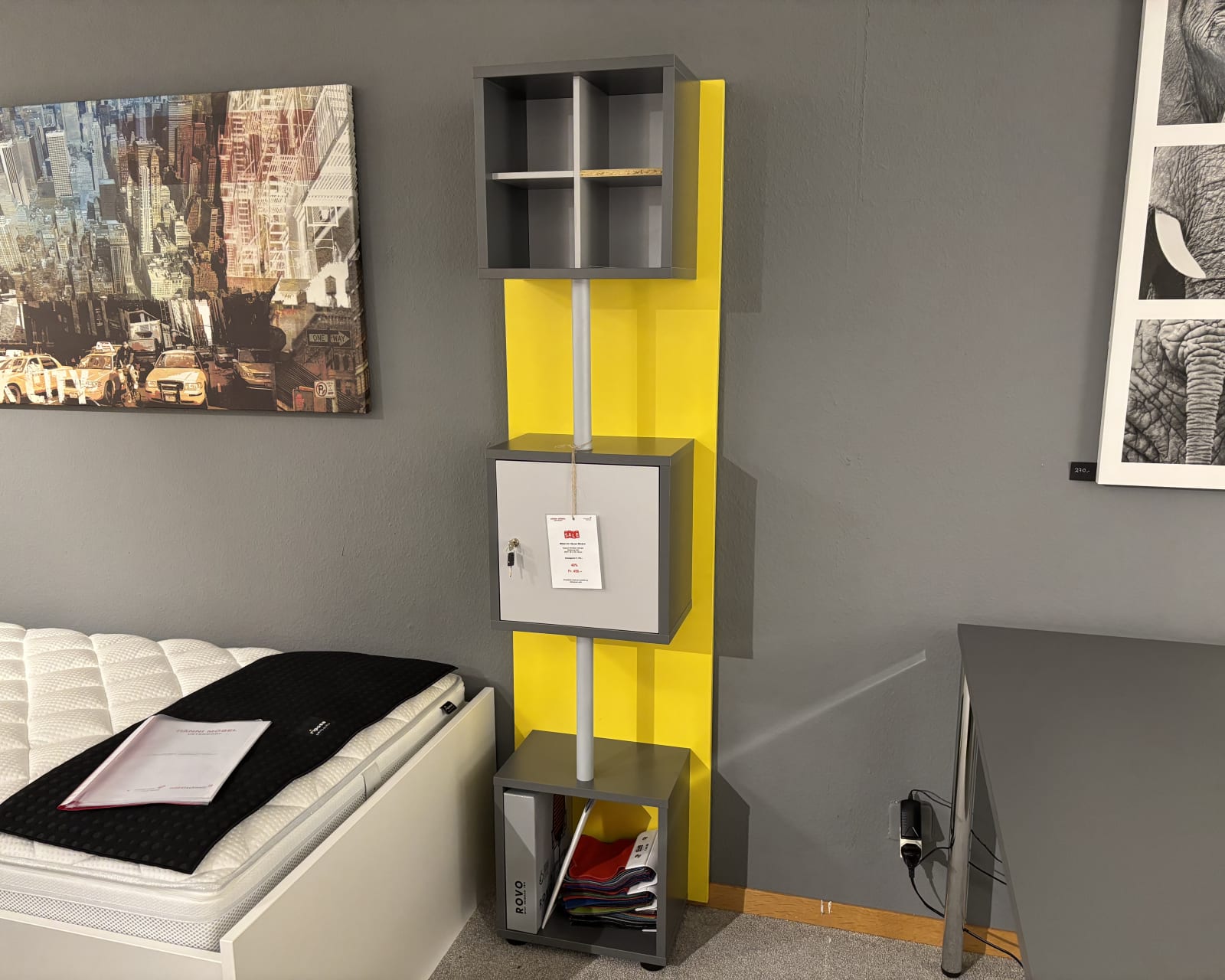 Das Büromöbel mit 3 Boxen Window, ein hohes Regal in Grau und Gelb, steht neben einem Bett mit weißer Matratze und schwarzer Decke.