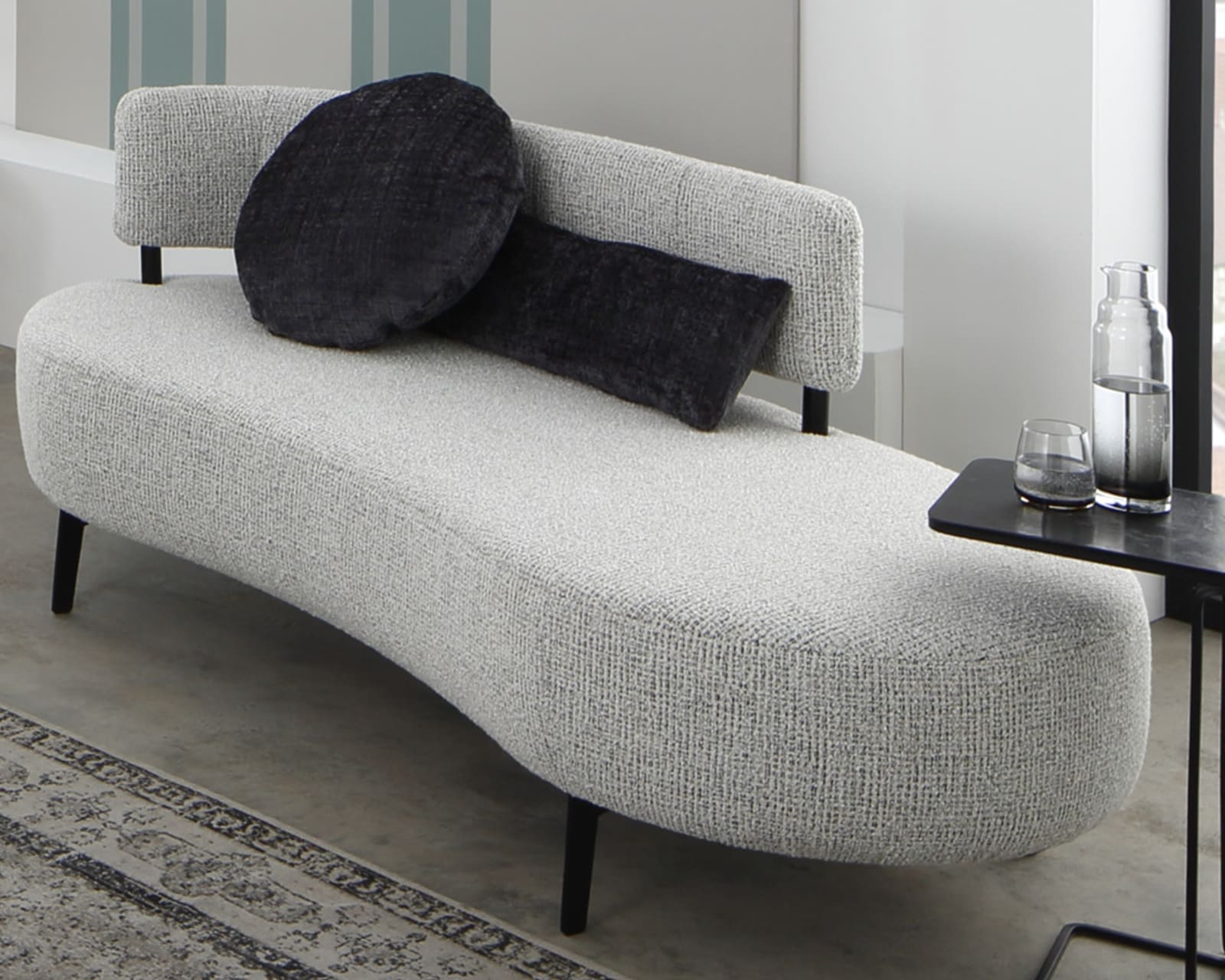 Ein geschwungenes graues Sofa von Ponsel Recamiere Organic mit zwei dunklen, runden Kissen steht neben einem schwarzen Beistelltisch, auf dem eine Glaskanne und ein Glas stehen.