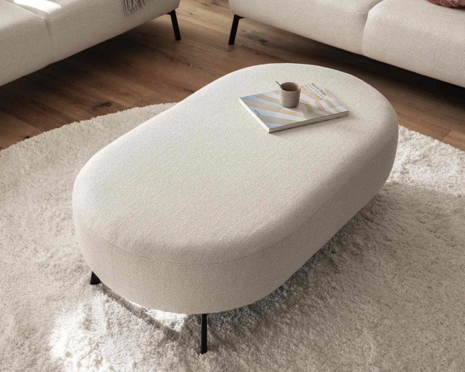 Das Candy Sofa Nimba dient als perfekte Kulisse für einen ovalen beigen Ottomanen, auf dem ein Buch und eine Tasse liegen, die alle auf einem runden beigen Teppich im Wohnzimmer ruhen.